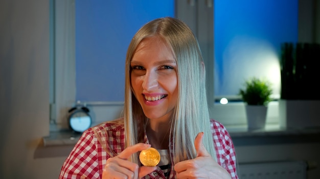 Vrolijke jonge blonde vrouw in vrijetijdskleding die 's nachts bij een donker raam zit met bitcoin en doi...