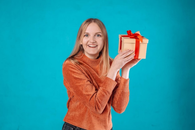 Vrolijke jonge blonde vrouw van 20 jaar draagt een groen spijkerjasje en houdt een rode geschenkdoos vast met een cadeaulint en probeert te raden wat erin zit geïsoleerd op een effen blauwe achtergrond studio portret
