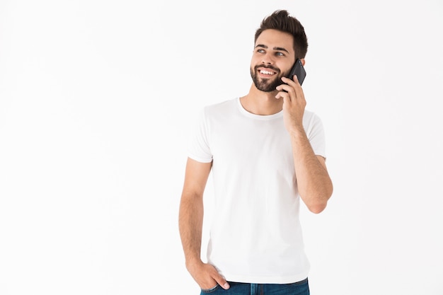 Vrolijke jonge brunette bebaarde man met een casual outfit die geïsoleerd over een witte muur staat en op mobiele telefoon praat