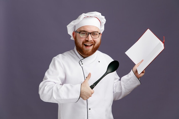 Vrolijke jonge chef-kok met een uniforme bril en een dop met een stevige lepel die een notitieblok toont en naar een camera kijkt met duim omhoog geïsoleerd op een paarse achtergrond