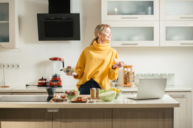 Vrolijke jonge dame die zich verrukt voelt en glimlacht terwijl ze in de keuken staat met salade en houten kom terwijl ze in de verte kijkt