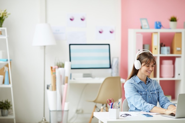 Vrolijke jonge dame in bedrade koptelefoon zittend aan tafel en het gebruik van laptop tijdens het werken met pleasure