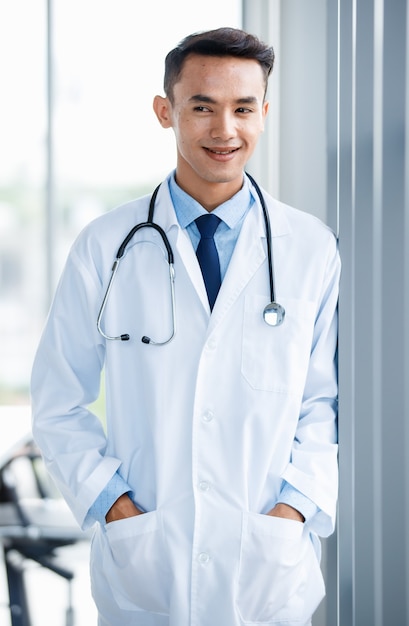 Vrolijke jonge en schattige Aziatische mannelijke arts in uniform en met een stethoscoop die in de kliniek staat en poseert met een positief en zelfverzekerd gebaar.