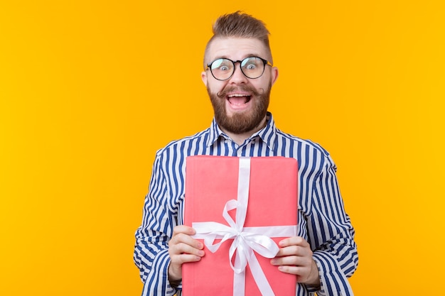 Vrolijke jonge hipster man met een snor in glazen knuffelt een rode geschenkdoos op een gele muur. Het concept van de vreugde van geschenken ...