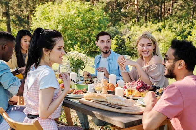 Vrolijke jonge interculturele mannen en vrouwen in vrijetijdskleding praten zittend aan tafel geserveerd met zelfgemaakte gerechten en vers fruit