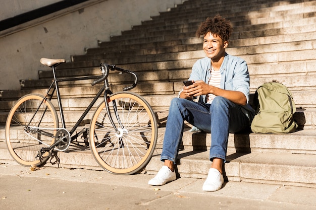 Vrolijke jonge kerel buiten zitten met fiets op straat op trappen met behulp van telefoon