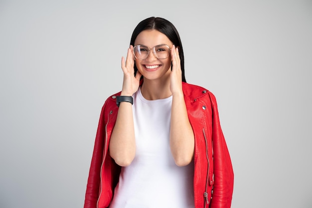 Vrolijke jonge knappe vrouw die haar bril corrigeert en binnenshuis poseert, terwijl ze breed glimlacht met haar witte rechte tanden, lacht om een goede grap, casual kleding draagt