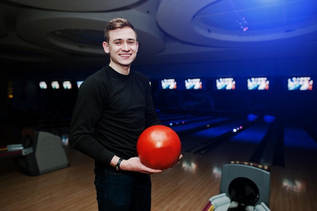 Vrolijke jonge man die een bowlingbal vasthoudt en naar de camera glimlacht terwijl hij tegen bowlingbanen staat met ultraviolet licht