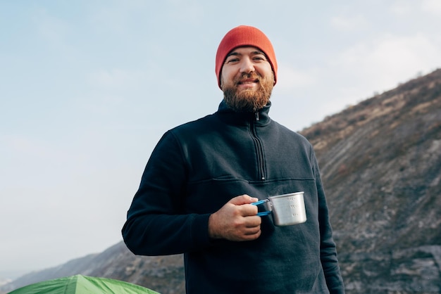 Vrolijke jonge man die thee of koffie drinkt in de bergen Reiziger man met baard met rode hoed die een mok thee in handen houdt na het wandelen in de bergen Reizen levensstijl en vakantie concept