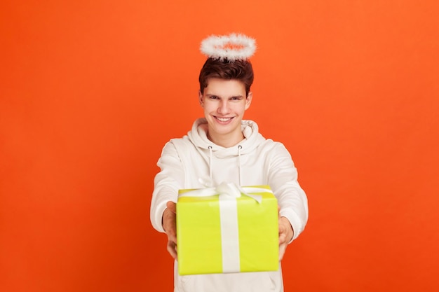 Vrolijke jonge man in casual sweatshirt met vriendelijke glimlach en halo overhead holding geschenkdoos liefdadigheid organisatie religie Indoor studio shot geïsoleerd op oranje achtergrond