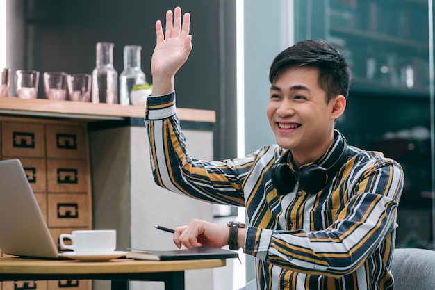 Foto vrolijke jonge man in een café die één hand opsteekt terwijl hij naar iemand in de verte zwaait