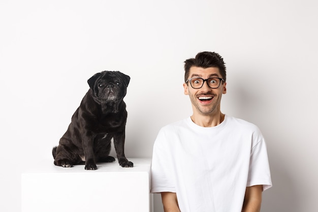 Vrolijke jonge man in glazen zitten in de buurt van schattige kleine hond en camera te kijken. Hondeneigenaar starend blij en verbaasd, wit