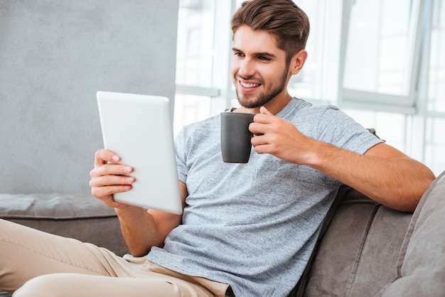 Vrolijke jonge man in grijs t-shirt zittend op de bank thuis. Communicatie via tablet en lachend tijdens het drinken van een kopje koffie.