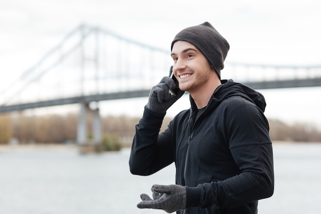 Vrolijke jonge man in hoed en handschoenen staan en praten op mobiele telefoon buitenshuis