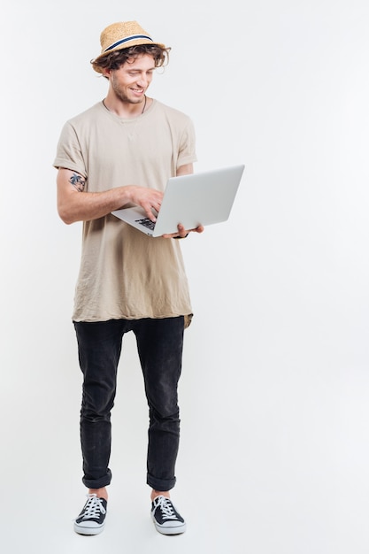 Vrolijke jonge man in hoed permanent en met behulp van laptop op witte achtergrond