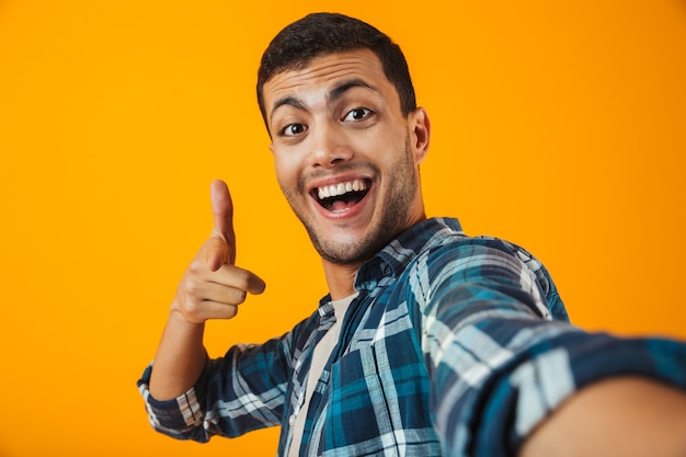 Vrolijke jonge man met een geruite hemd staande geïsoleerd over oranje muur, een selfie nemen