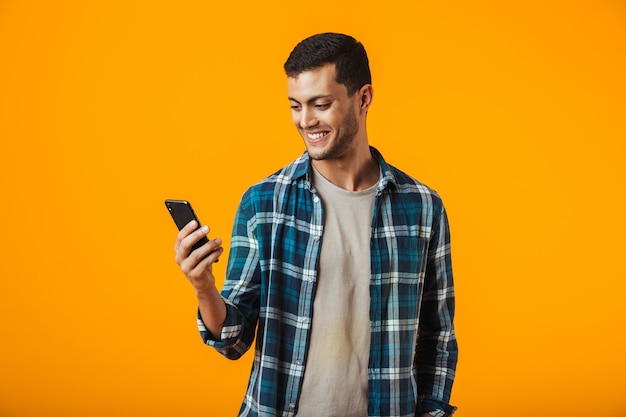 Vrolijke jonge man met geruite hemd staande geïsoleerd over oranje muur, met behulp van mobiele telefoon