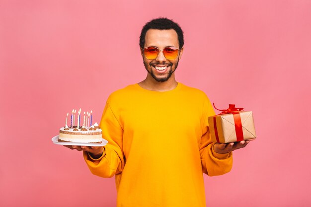 Vrolijke jonge man met geschenkdoos blaast kaarsjes op een verjaardagstaart geïsoleerd op roze.