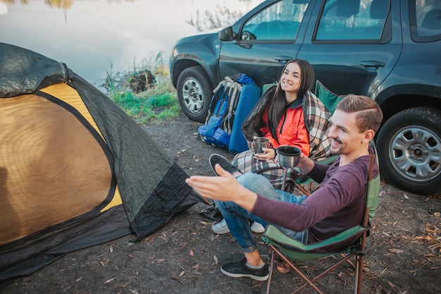 Vrolijke jonge man op zoek naar links en hand vast te houden. Vrouw kijkt in dezelfde richting. Ze hebben thermocups in handen. Mensen zitten bij tent met auto.