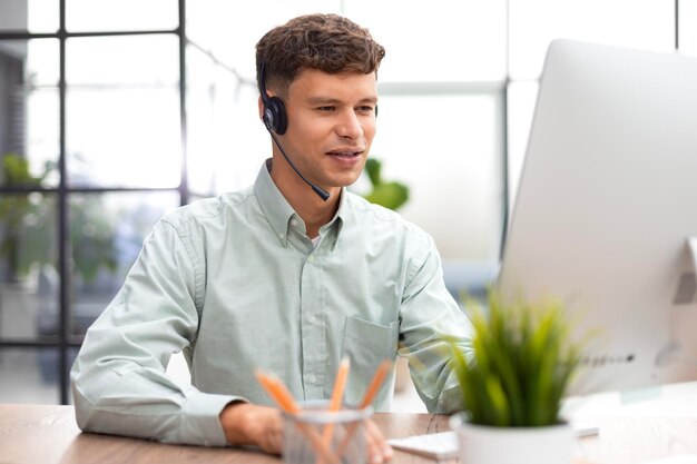 Vrolijke jonge mannelijke ondersteuningstelefoonoperator in headset op de werkplek tijdens het gebruik van computerhulpservice en klantadvies callcenterconcept