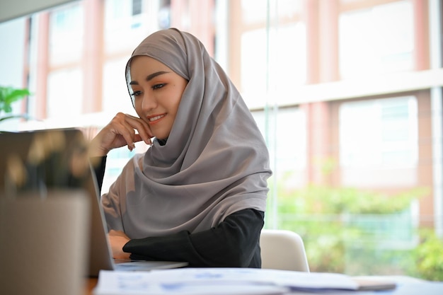 Vrolijke jonge moslimvrouw die hijab draagt op haar bureau en naar het scherm van de laptop kijkt