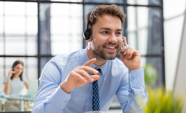 Vrolijke jonge ondersteuning telefoon mannelijke operator in headset op werkplek met collega's op de achtergrond tijdens het gebruik van computer help service en client consulting callcenter concept