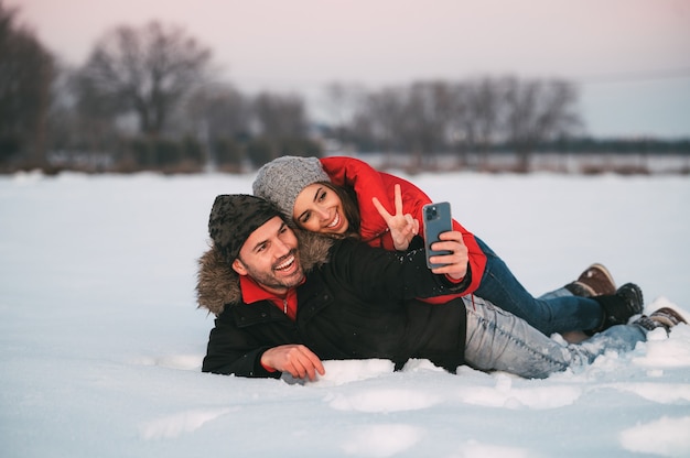 Vrolijke jonge paar in warme kleren liggen samen in de sneeuw en nemen selfie op smartphone terwijl ze plezier hebben in de winterlandschap