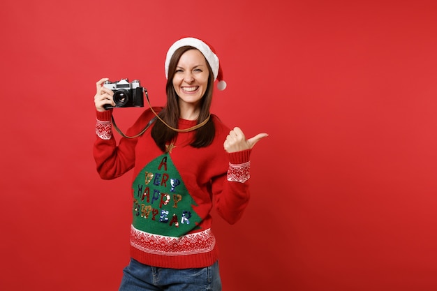 Vrolijke jonge Santa meisje in kerstmuts duim opzij wijzen, retro vintage fotocamera geïsoleerd op rode achtergrond te houden. Gelukkig Nieuwjaar 2019 viering vakantie partij concept. Bespotten kopie ruimte.