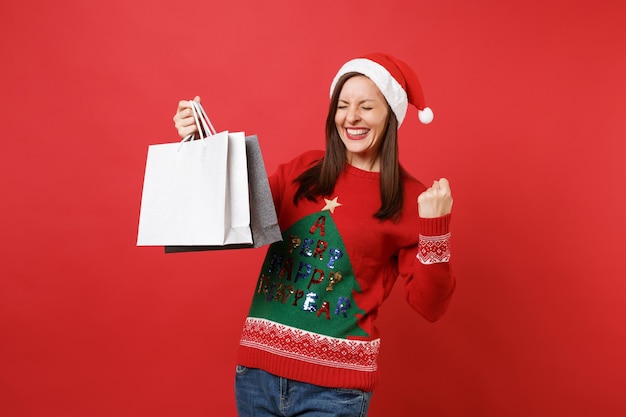 Vrolijke jonge Santa meisje met gesloten ogen winnaar gebaar doen, pakketten tassen met aankopen houden na het winkelen geïsoleerd op rode achtergrond. Gelukkig Nieuwjaar 2019 viering vakantie partij concept.