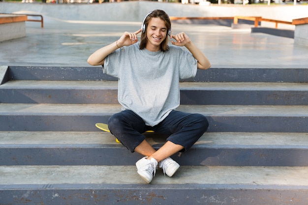 Vrolijke jonge tiener tijd doorbrengen in het skatepark, luisteren naar muziek met een koptelefoon, zittend op het skateboard