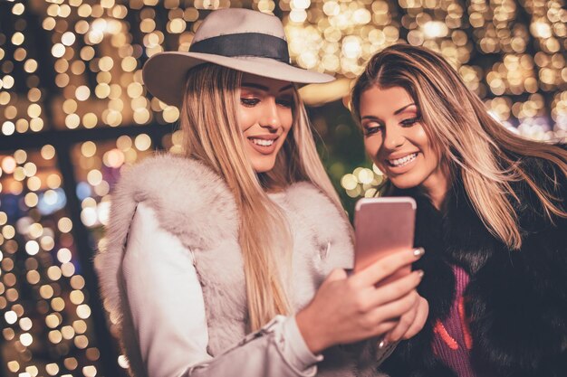 Vrolijke jonge vriendinnen hebben plezier in de straat van de stad tijdens de kerstnacht. Ze surfen op het sociale netwerk op de smartphone en praten iets grappigs.