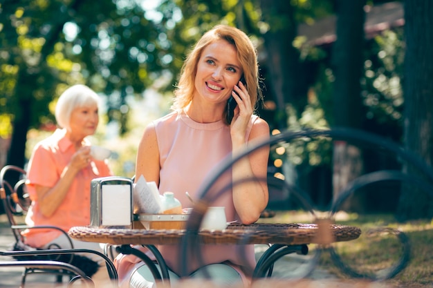 Vrolijke jonge vrouw die buiten aan de cafétafel zit en glimlacht tijdens een telefoongesprek