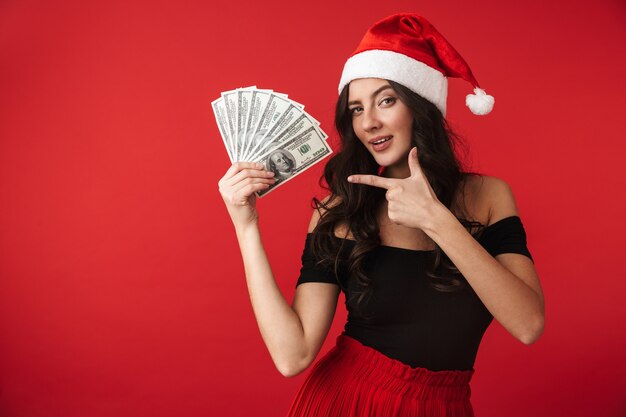 Vrolijke jonge vrouw die Kerstmishoed draagt die zich geïsoleerd over rood bevindt, die geldbankbiljetten toont