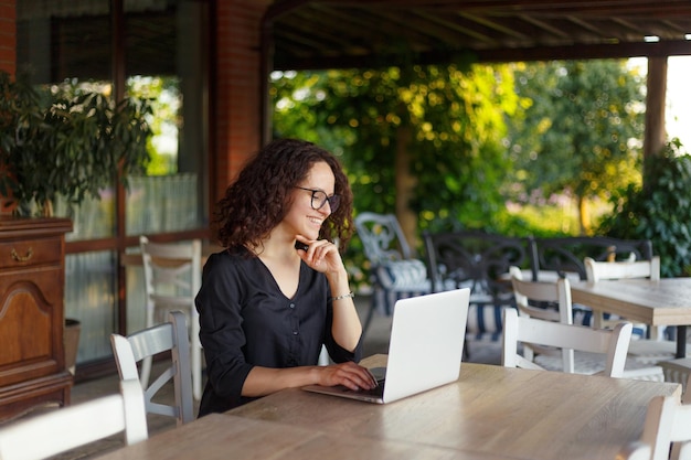 Vrolijke jonge vrouw die op laptop werkt terwijl ze op het terras zit