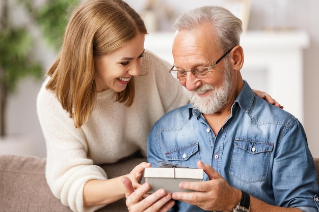 Vrolijke jonge vrouw die senior man knuffelt terwijl ze op de bank zit en de huidige doos opent tijdens de vakantieviering thuis