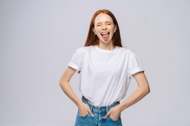 Vrolijke jonge vrouw die t-shirt en denimbroek draagt die tong op geïsoleerde witte achtergrond toont
