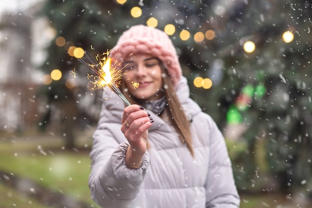 Vrolijke jonge vrouw draagt een roze gebreide muts en jas die plezier heeft met sterretjes op straat bij de kerstboom tijdens de sneeuwval