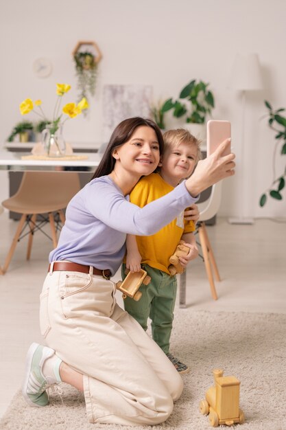 Vrolijke jonge vrouw en haar schattige zoontje smartphone camera kijken tijdens het maken van selfie thuis tijdens zelfisolatie