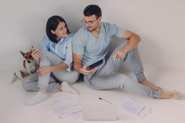 Vrolijke jonge vrouw gebruikt bankieren app op haar mobiele telefoon zijn man toont cijfers op rekenmachine omringd met papieren veel werk economische problemen oplossen samen iets bespreken