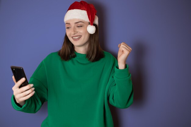 Vrolijke jonge vrouw gekleed in een groene trui en kerstmuts met een mobiele telefoon in haar hand die een winnaarsgebaar maakt geïsoleerd op een groene achtergrond Gelukkig nieuwjaar 2023 vakantieconcept