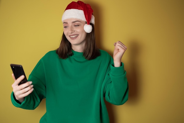 Vrolijke jonge vrouw gekleed in een groene trui en kerstmuts met een mobiele telefoon in haar hand die een winnaarsgebaar maakt geïsoleerd op een groene achtergrond Gelukkig nieuwjaar 2023 vakantieconcept