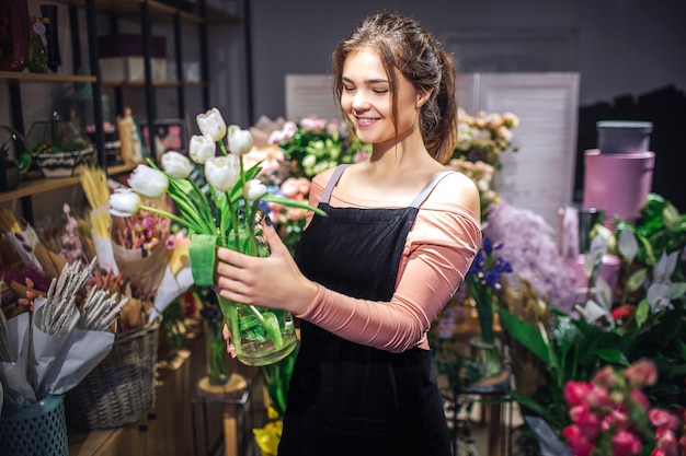 Vrolijke jonge vrouw houden glazen bloemenvaas met witte tulpen. Ze lacht. Bloemist staan in bloemenwinkel.