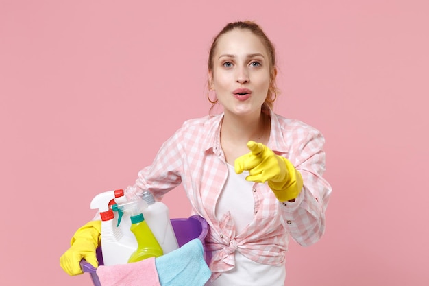 Vrolijke jonge vrouw huisvrouw in rubberen handschoenen houden bekken met wasmiddelflessen wassen reinigingsmiddelen huishoudelijk werk geïsoleerd op roze achtergrond. Huishoudelijk concept. Wijzende wijsvinger op camera.