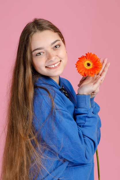 Vrolijke jonge vrouw in blauwe hoodie die zacht oranje bloem vasthoudt terwijl ze naar de camera lacht