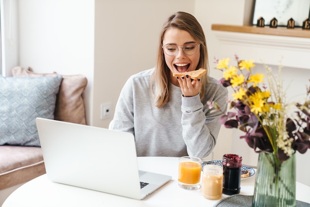 Vrolijke jonge vrouw in brillen die toast eet terwijl ze een laptop gebruikt in de woonkamer