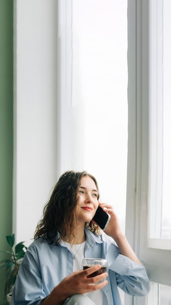 Vrolijke jonge vrouw in casual kleding chatten en flirten met haar vriendje aan de telefoon zittend op een vensterbank met een kopje zwarte thee