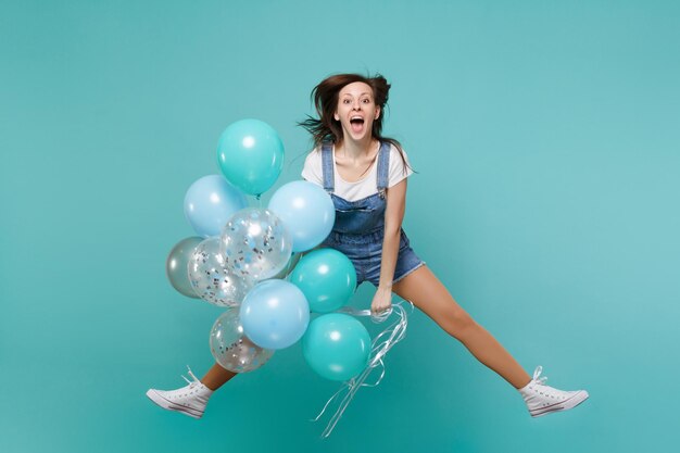 Vrolijke jonge vrouw in denim kleding houdt mond open, springt hoog, viert, houdt kleurrijke luchtballonnen geïsoleerd op blauwe turkooizen achtergrond. Vakantie verjaardagsfeestje, mensen emoties concept.