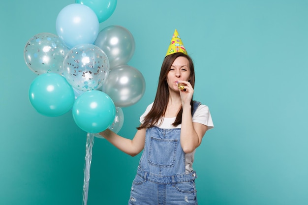 Vrolijke jonge vrouw in denim kleding, verjaardag hoed waait in pijp, vieren, kleurrijke lucht ballonnen geïsoleerd op blauwe turkooizen achtergrond te houden. Vakantie verjaardagsfeestje, mensen emoties concept.