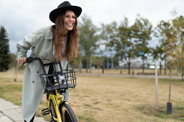Vrolijke jonge vrouw in een grijze jas en zwarte hoed fietst met een glimlach