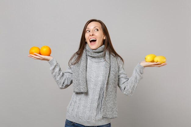 Vrolijke jonge vrouw in grijze trui, sjaal opzoeken, met citroenen sinaasappelen geïsoleerd op een grijze achtergrond. Gezonde mode-levensstijl, oprechte emoties van mensen, concept van het koude seizoen. Bespotten kopie ruimte.